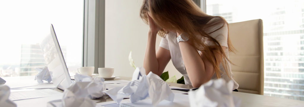 Upset woman at desk holding her head in her hands with wads of paper everywhere.